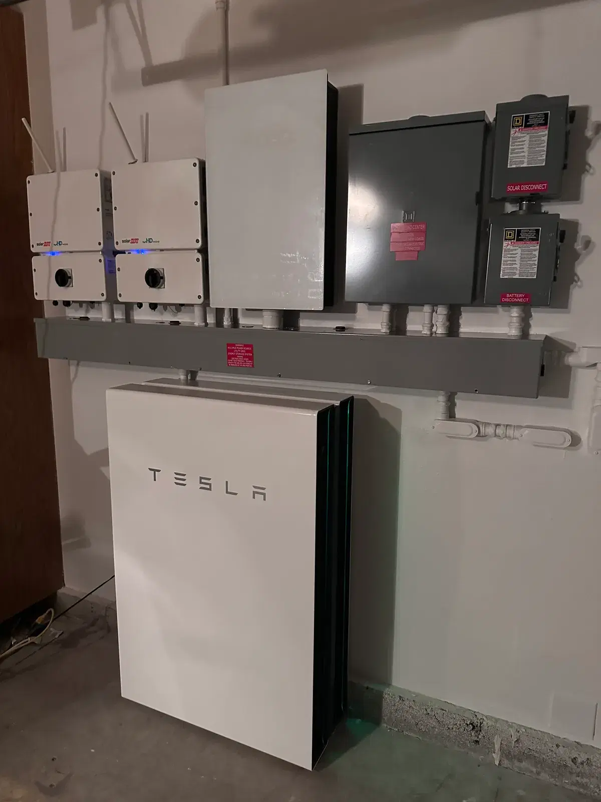 A wall-mounted Tesla Powerwall battery beneath a Tesla Backup Gateway, two SolarEdge inverters, and several gray electrical disconnect boxes labeled solar disconnect and battery disconnect.