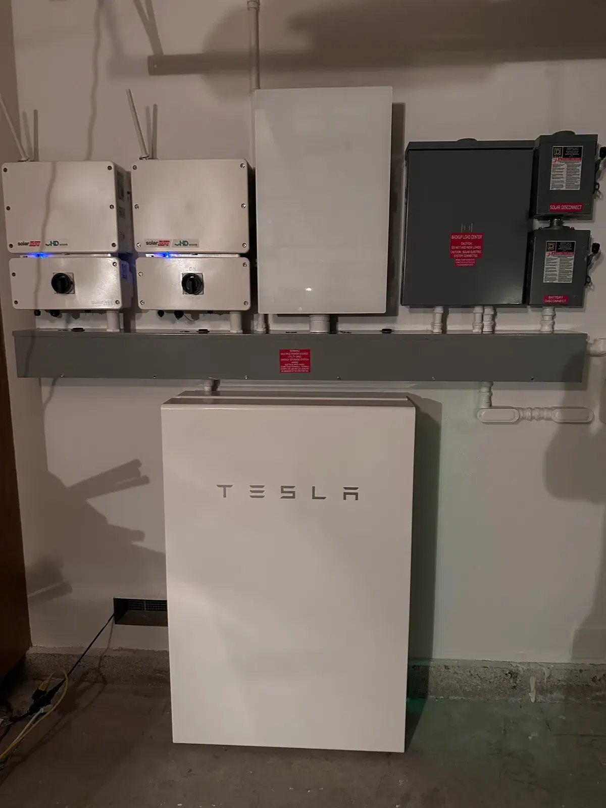 A white Tesla Powerwall battery mounted on a garage wall beneath two SolarEdge HD-Wave inverters, a backup load center, and solar and battery disconnect switches.