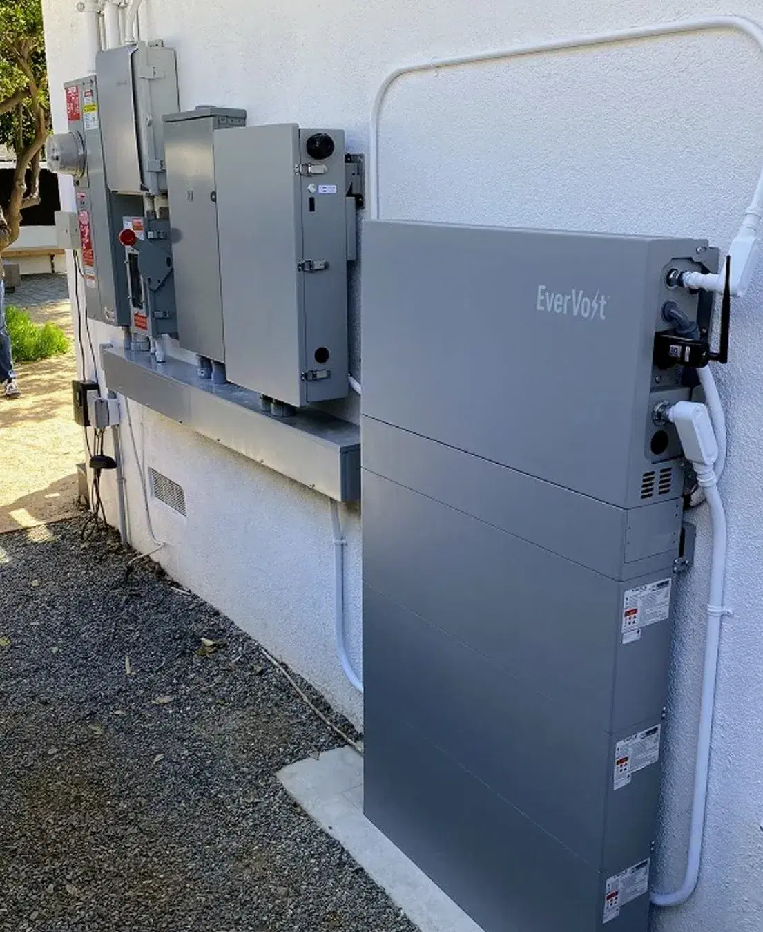 A tall gray EverVolt battery cabinet mounted on a stucco exterior wall next to electrical sub-panels, disconnects, and white conduit.