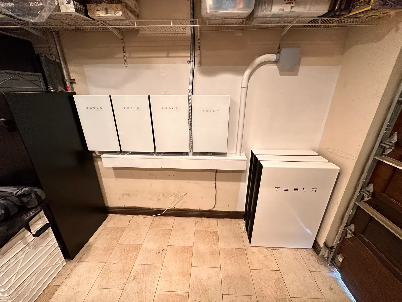 A garage interior with four wall-mounted Tesla Powerwall batteries on a ledge and one floor-standing unit beside them.