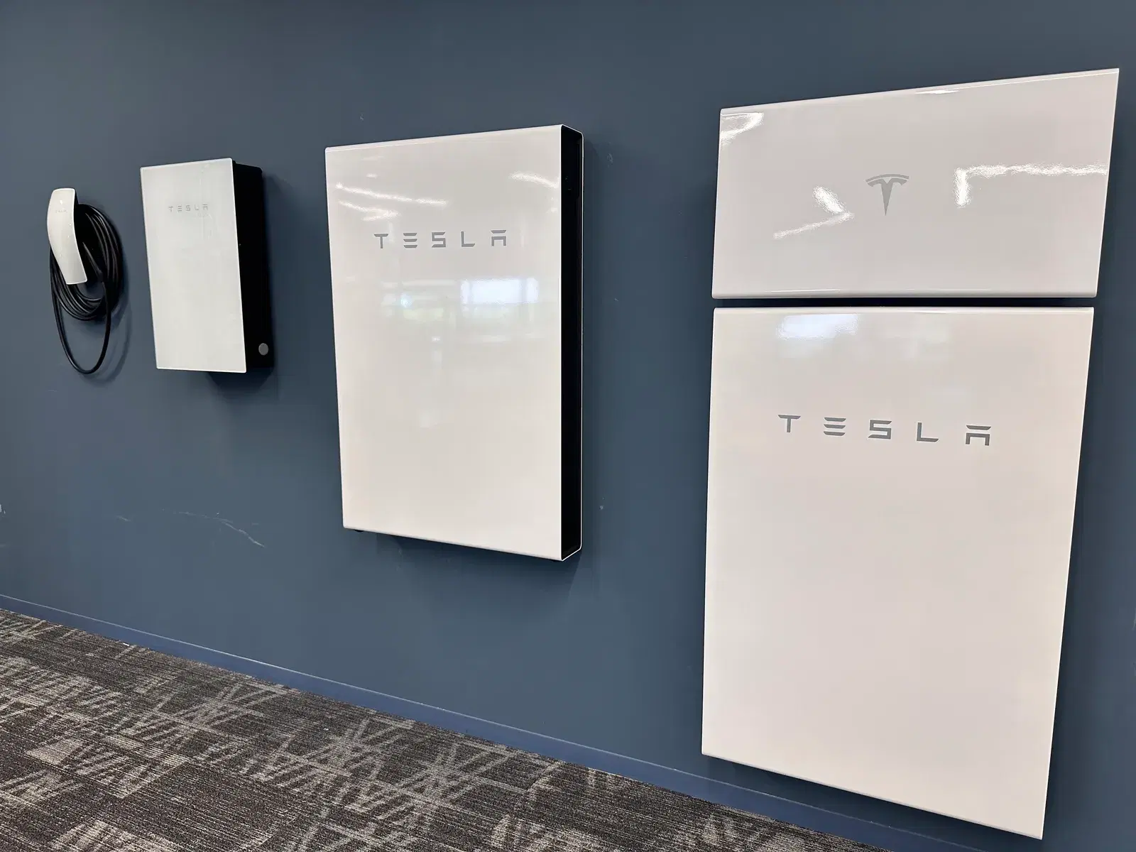 Interior wall display of Tesla home energy equipment including an EV Wall Connector with coiled cable, a Backup Gateway, and two white Powerwall batteries.