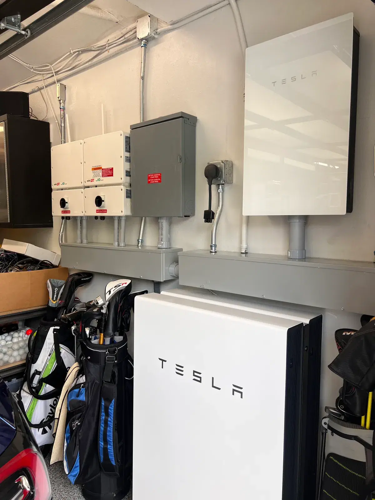 Two white Tesla Powerwall batteries mounted on a garage wall with electrical sub-panels, disconnects, and gray conduit, golf bags stored nearby.