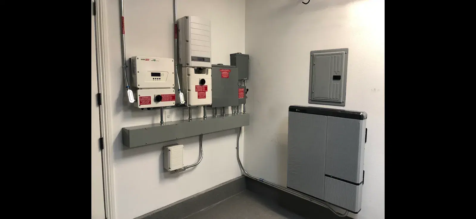 Interior utility room with a gray LG home battery on the right and a SolarEdge inverter with disconnect switches mounted on the wall.