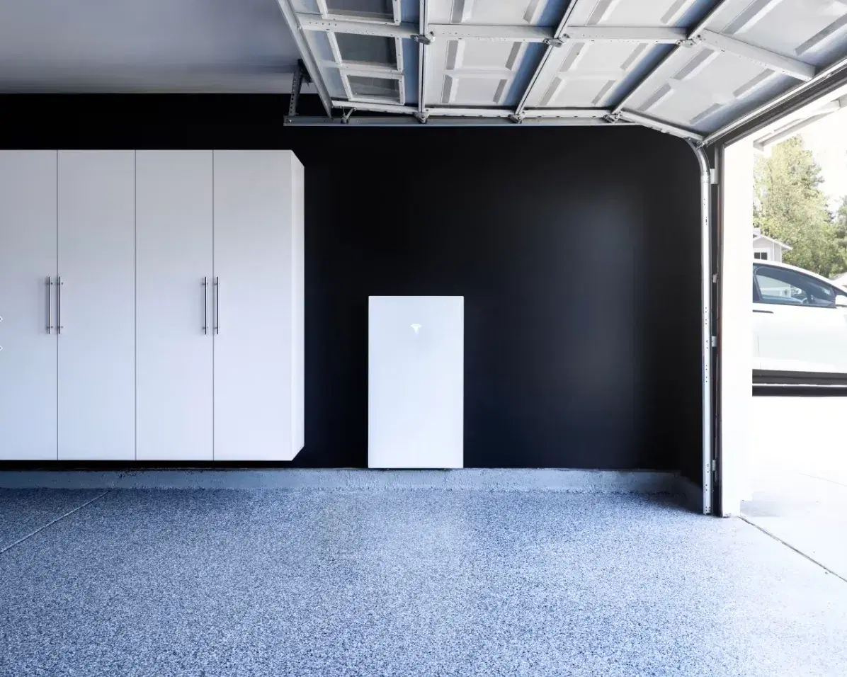 White Tesla Powerwall battery mounted on a black wall inside a clean garage with a Tesla car in the driveway.
