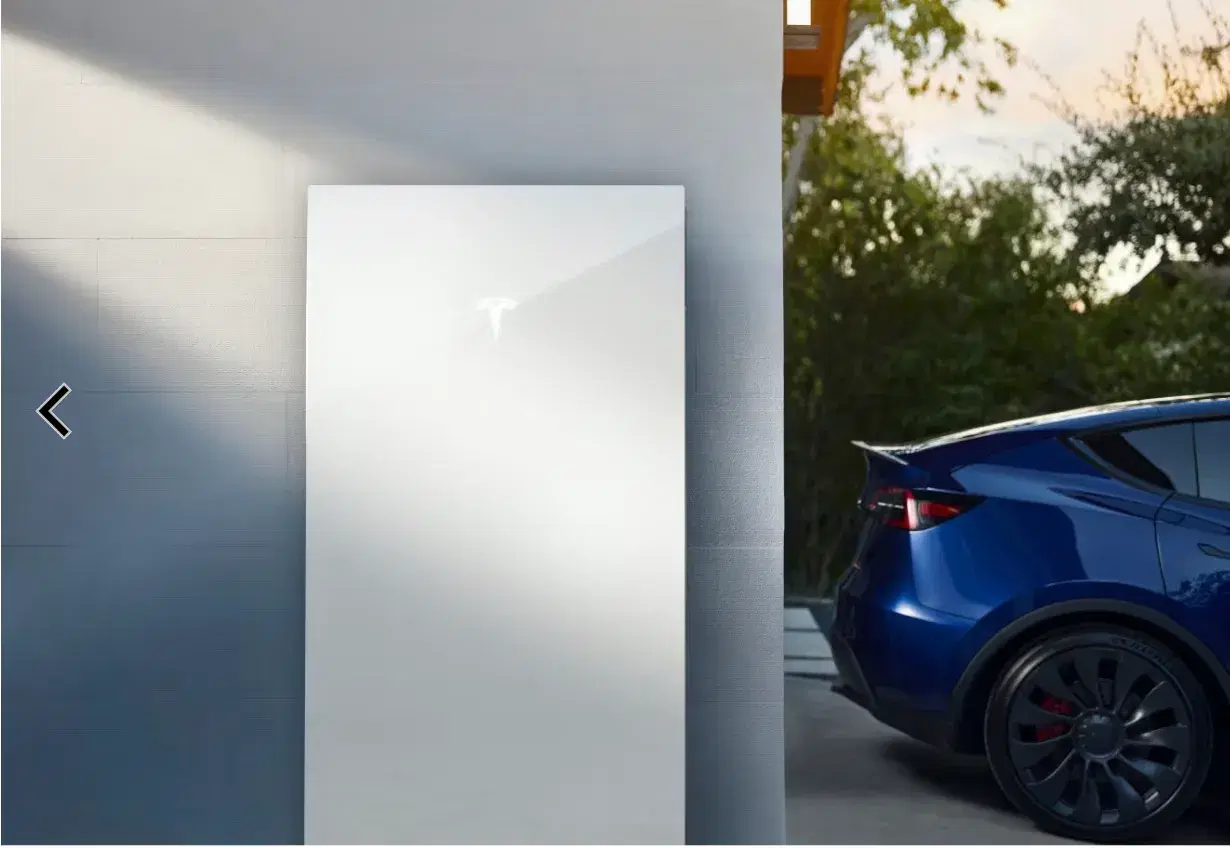 White Tesla Powerwall mounted on an outdoor wall at dusk with a blue Tesla Model Y parked alongside.