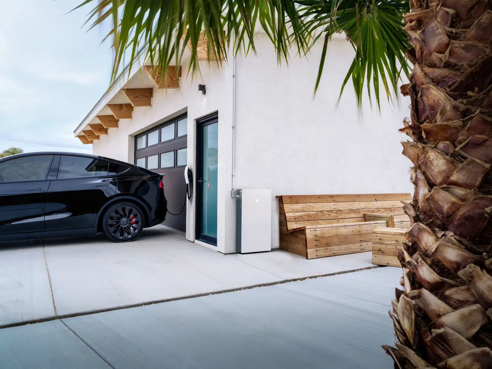 White Tesla Powerwall mounted on a home's exterior wall next to a garage, with a black electric vehicle charging nearby.