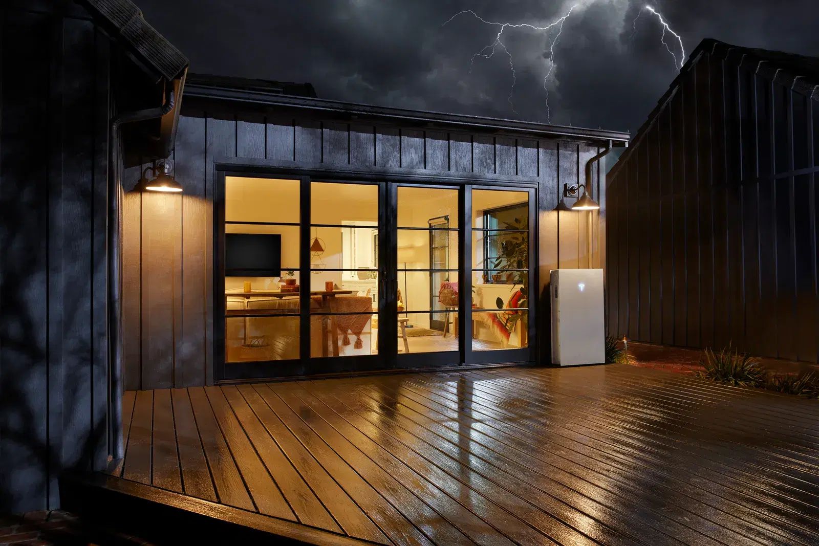 A wall-mounted Tesla Powerwall on a wooden patio at night, with lightning overhead and a brightly lit living room visible through glass doors.