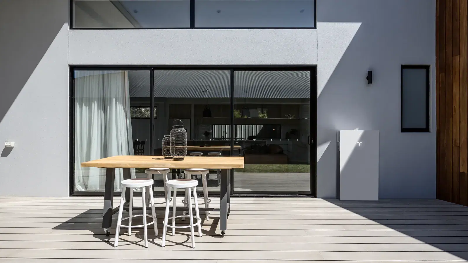 An outdoor dining table on a sunlit deck beside a contemporary white home, with a Tesla Powerwall mounted on the exterior wall.