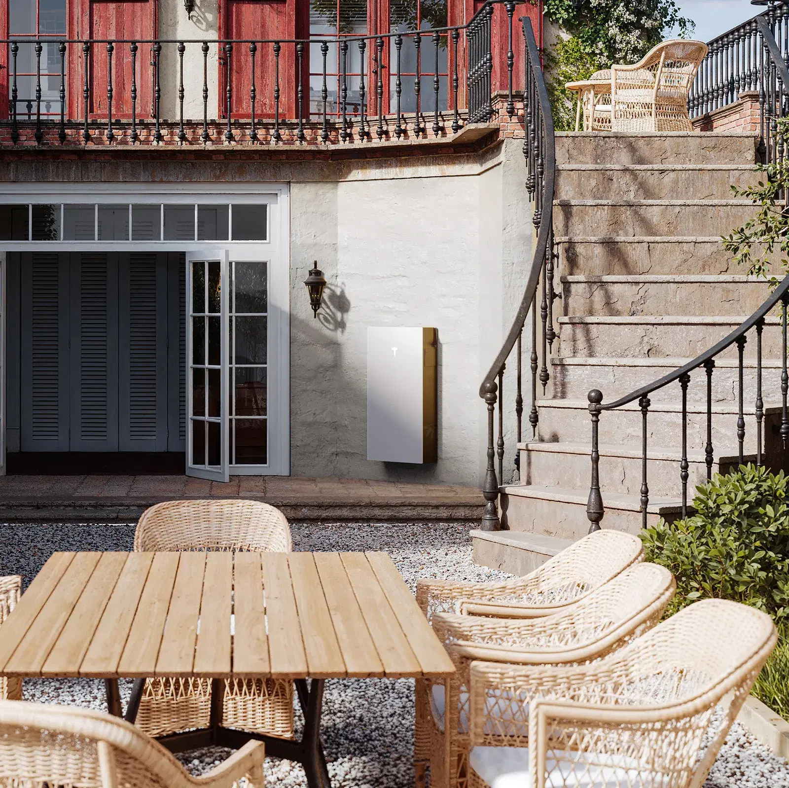 A white Tesla Powerwall mounted on a stucco wall in an elegant courtyard with woven dining chairs and a stone staircase.