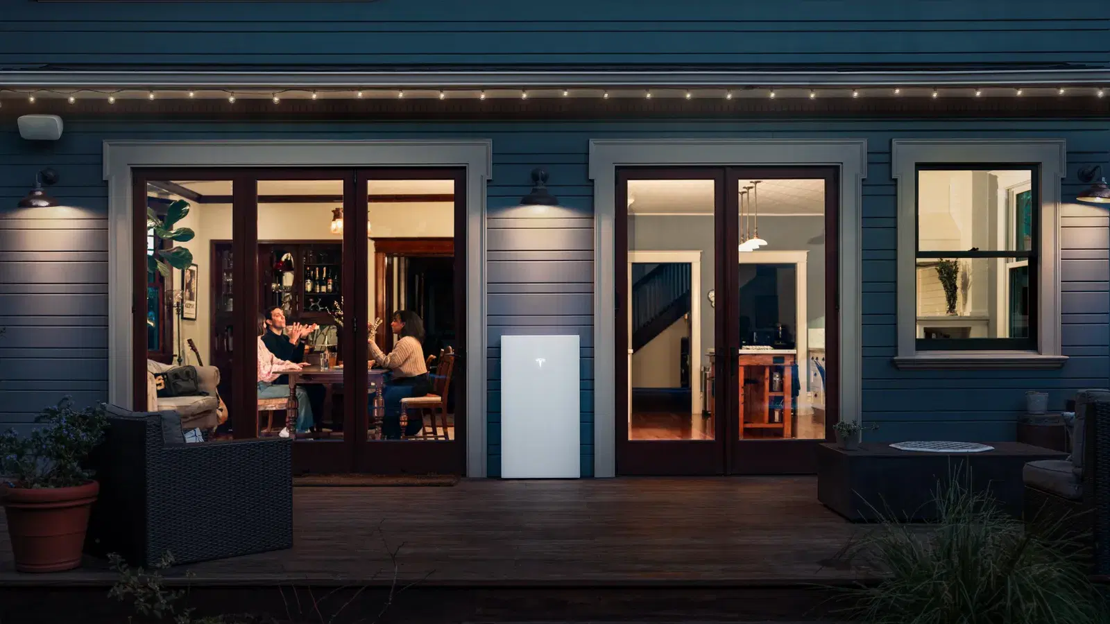 A white Tesla Powerwall mounted on a home's exterior wall at dusk, with people dining visible through open French doors.