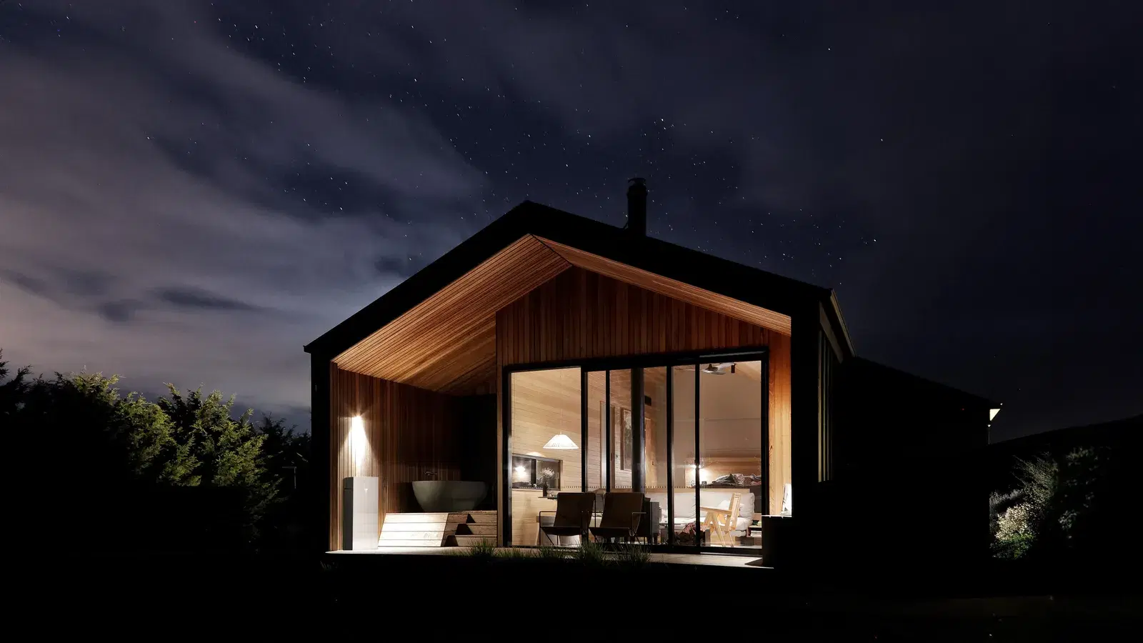 A modern timber cabin lit from within at night beneath a starry sky, with a Tesla Powerwall under the porch overhang.