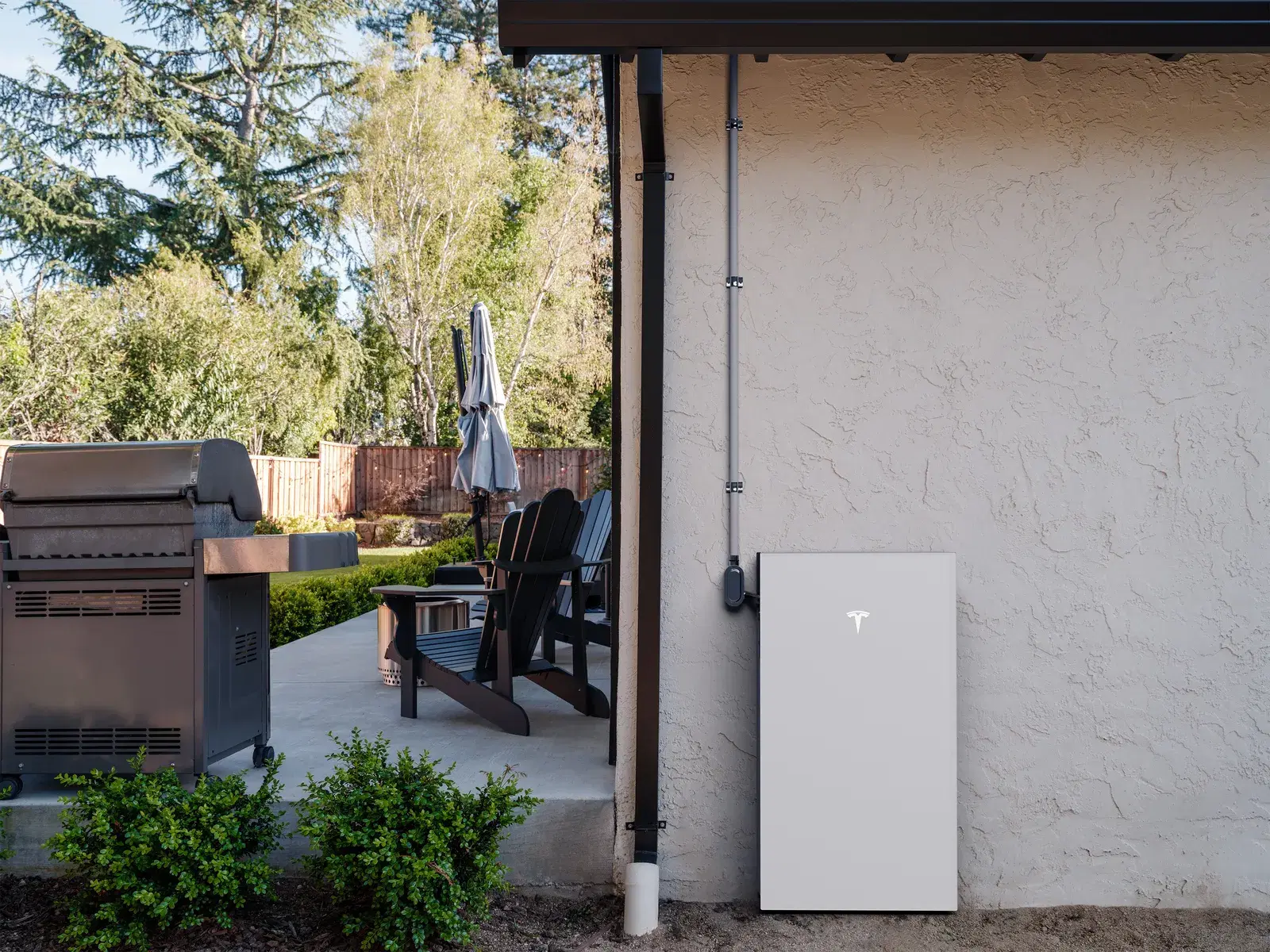 A white Tesla Powerwall with conduit mounted on a stucco exterior wall beside a backyard patio with a grill.