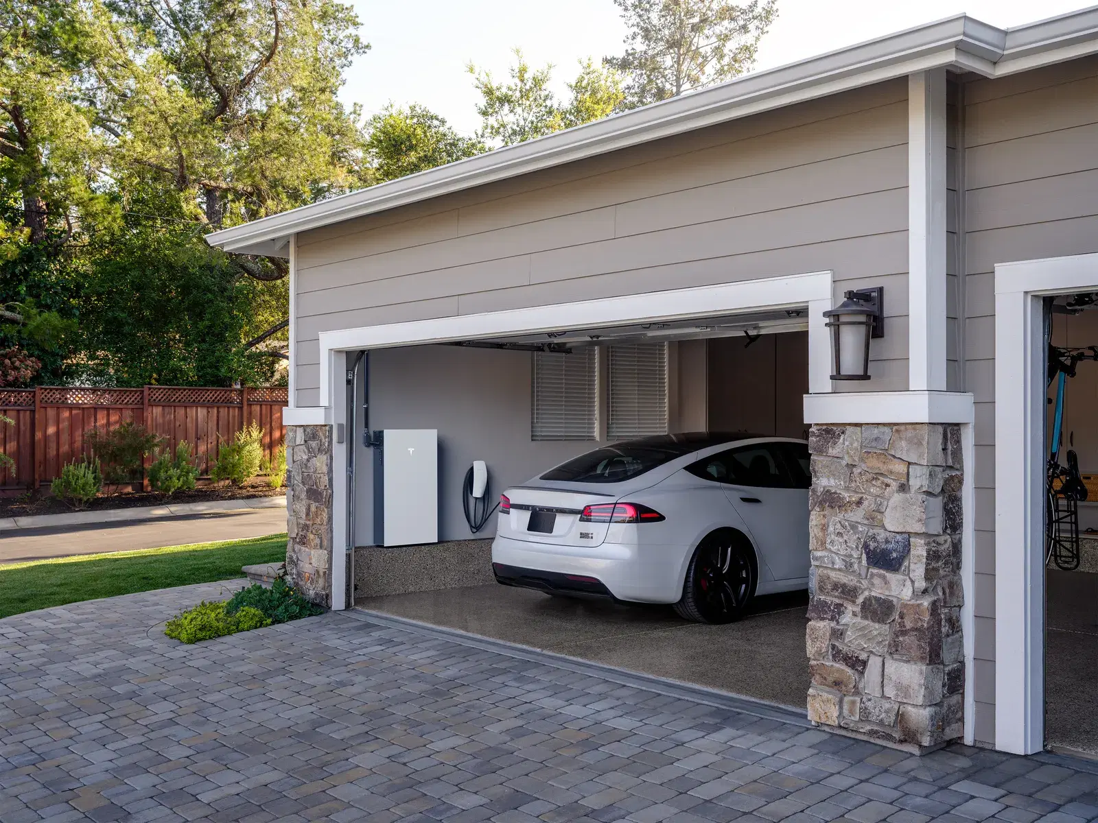 A white Tesla Powerwall and Wall Connector mounted inside an open garage with a white Tesla sedan parked beside them.