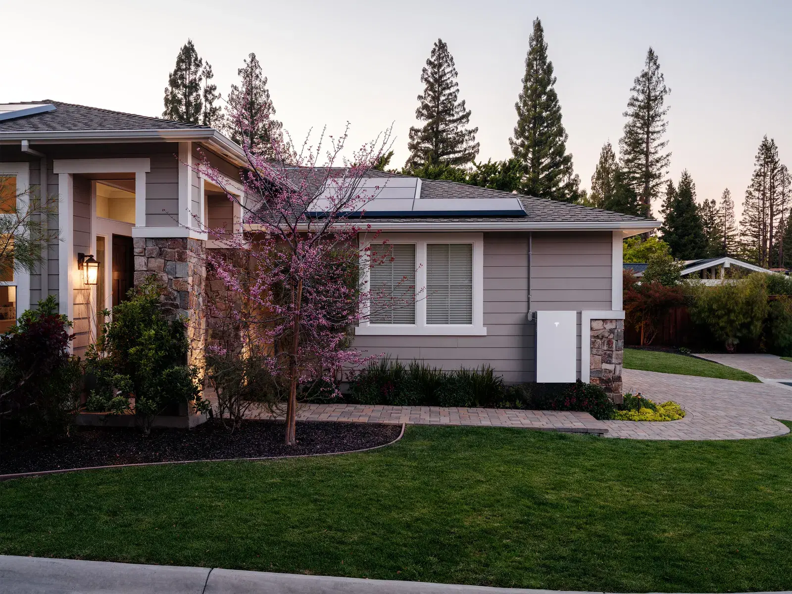 A Tesla Powerwall mounted on the side wall of a suburban home at twilight with a flowering pink tree in the foreground.