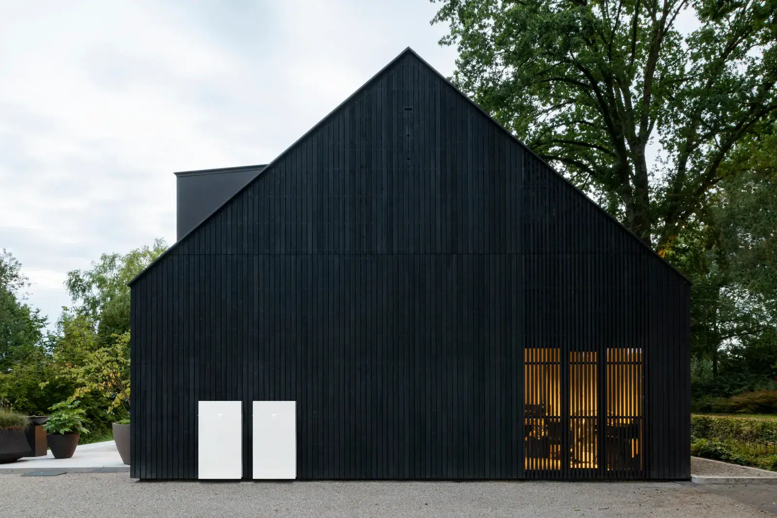 Two white Tesla Powerwall batteries mounted on the dark vertical-slat facade of a modern barn-style house.