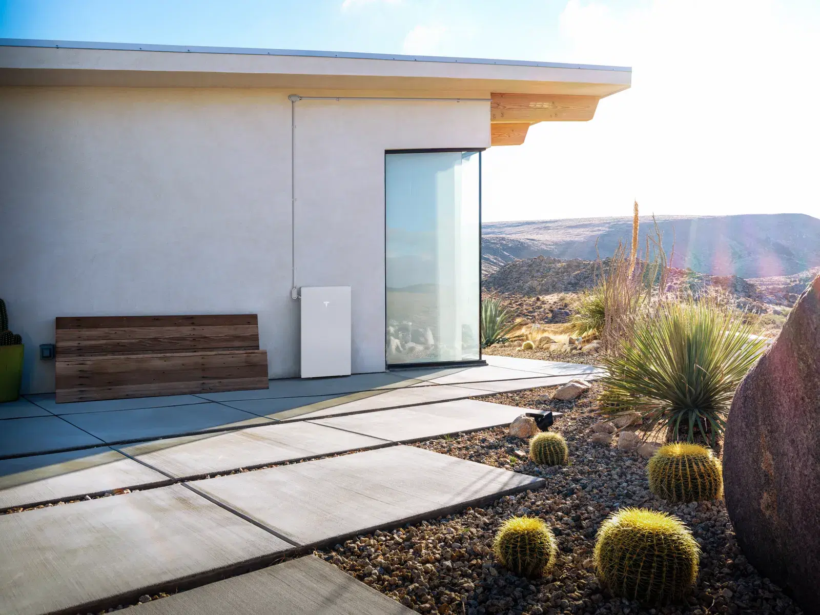 A white Tesla Powerwall on the exterior wall of a desert home with cactus landscaping and canyon views in the background.