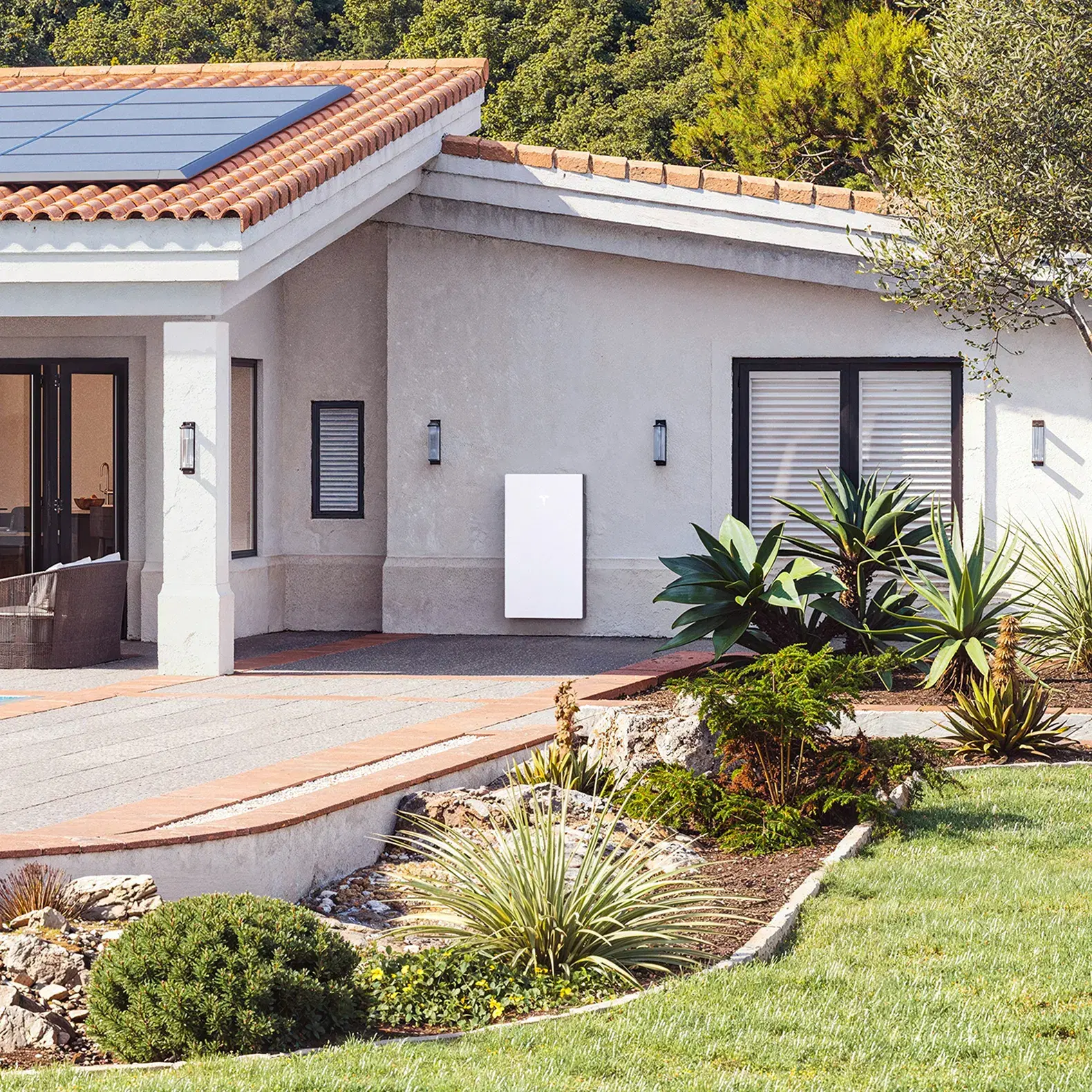 A Tesla Powerwall mounted on a white Mediterranean-style home with terracotta tile roof, solar panels, and drought-tolerant landscaping.