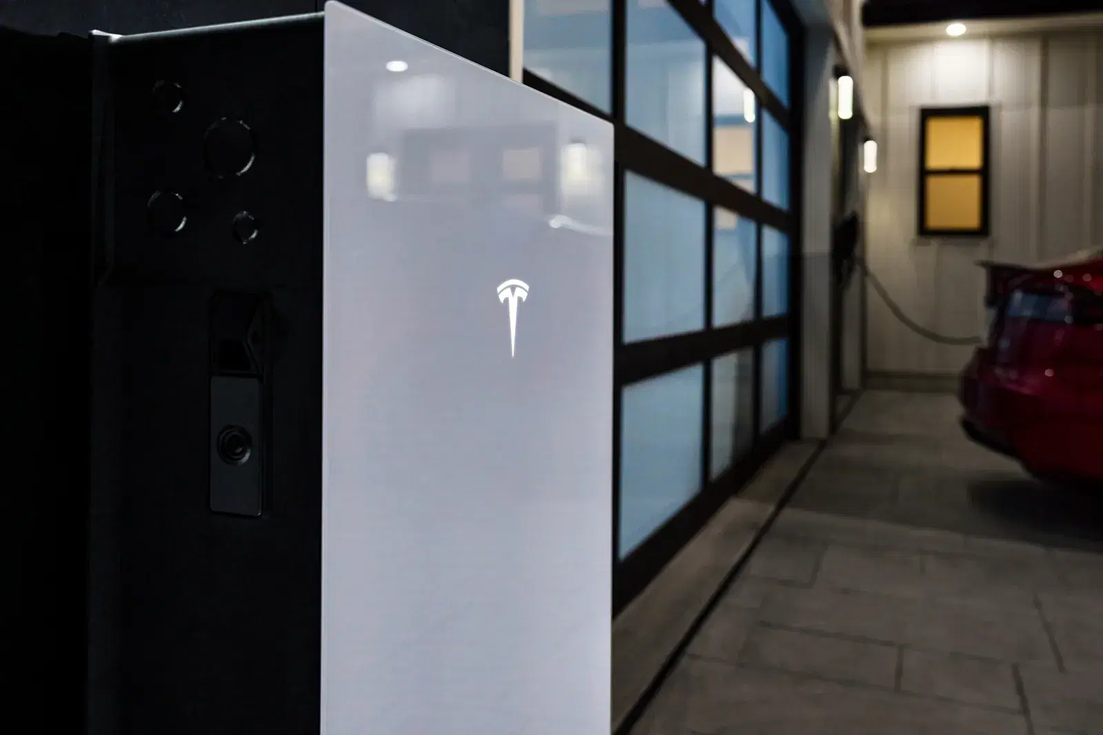 A detailed close-up of a Tesla Powerwall with illuminated logo, a red Tesla parked in a garage in the soft-focus background.