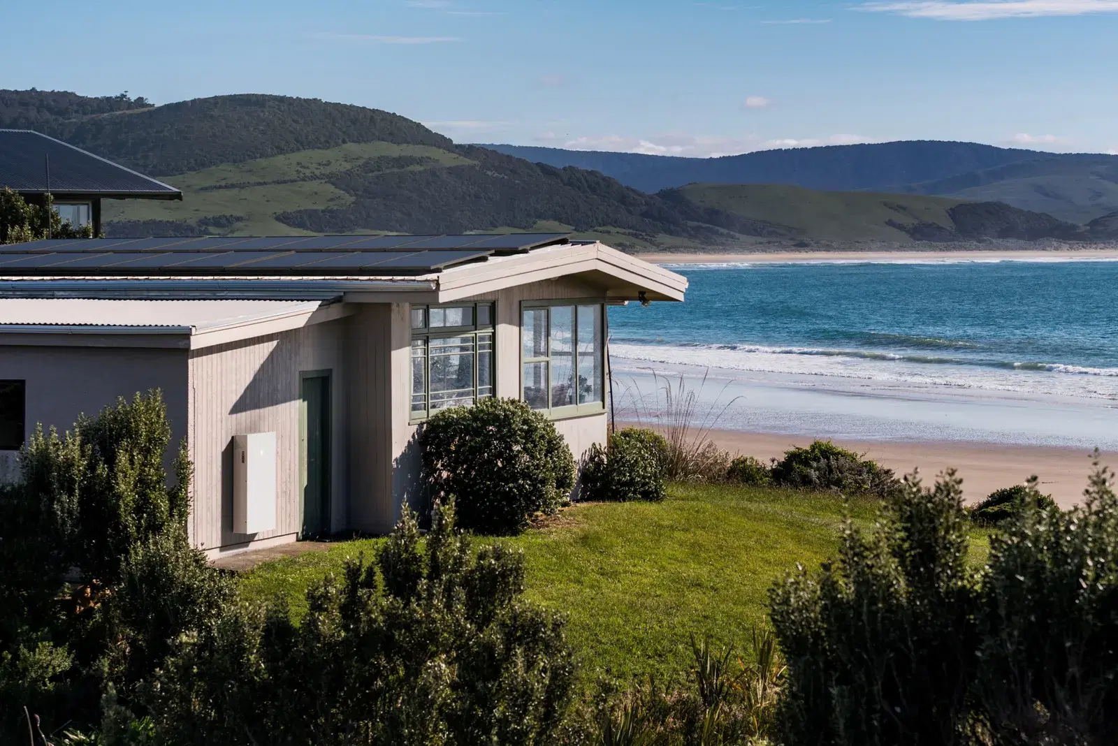 A beachfront home overlooking the sea with a white Tesla Powerwall mounted on its side wall and panels along the roof ridge.