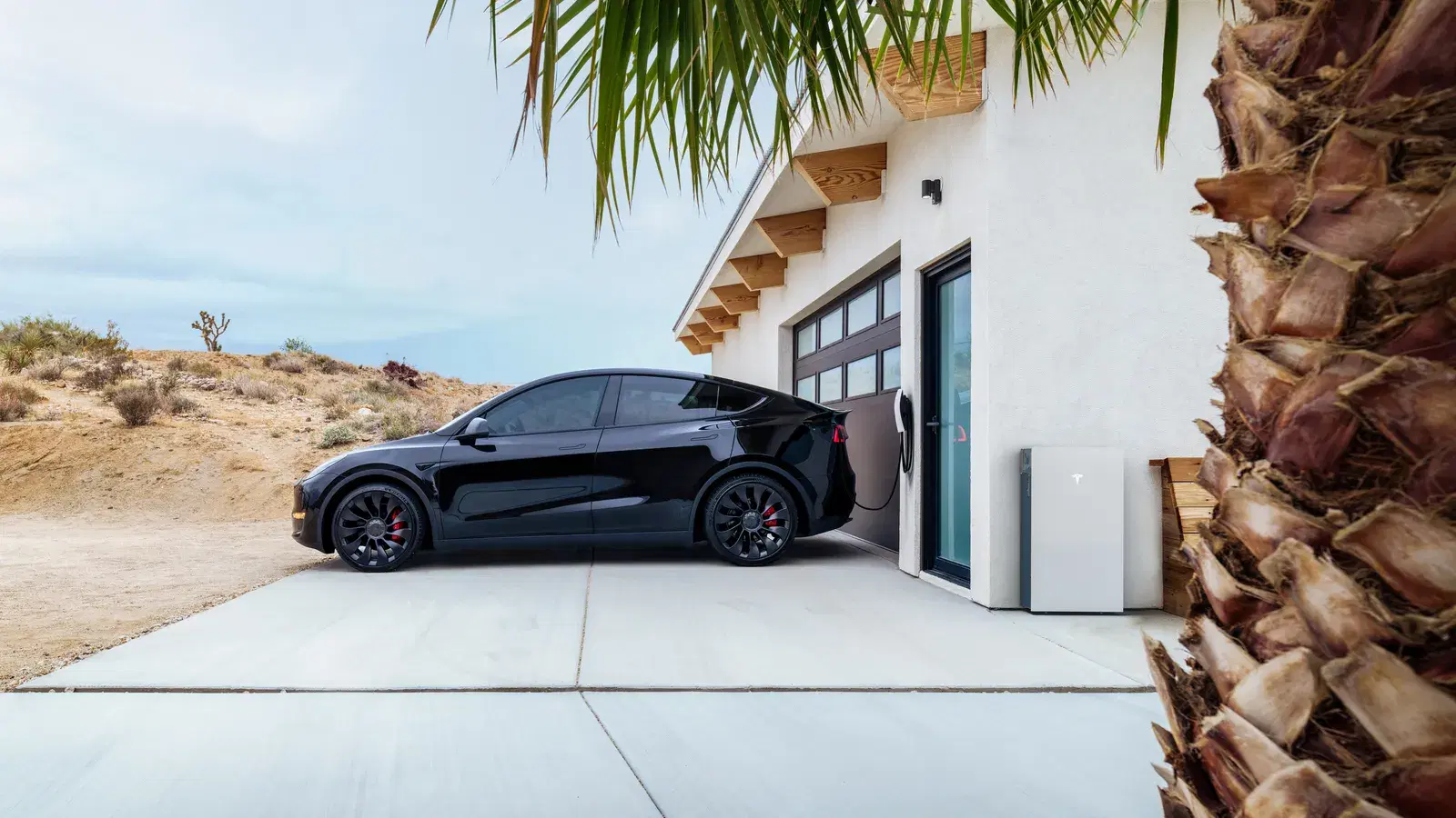 Black Tesla SUV parked at a desert home, plugged into a wall charger next to a white Tesla Powerwall on the garage wall.