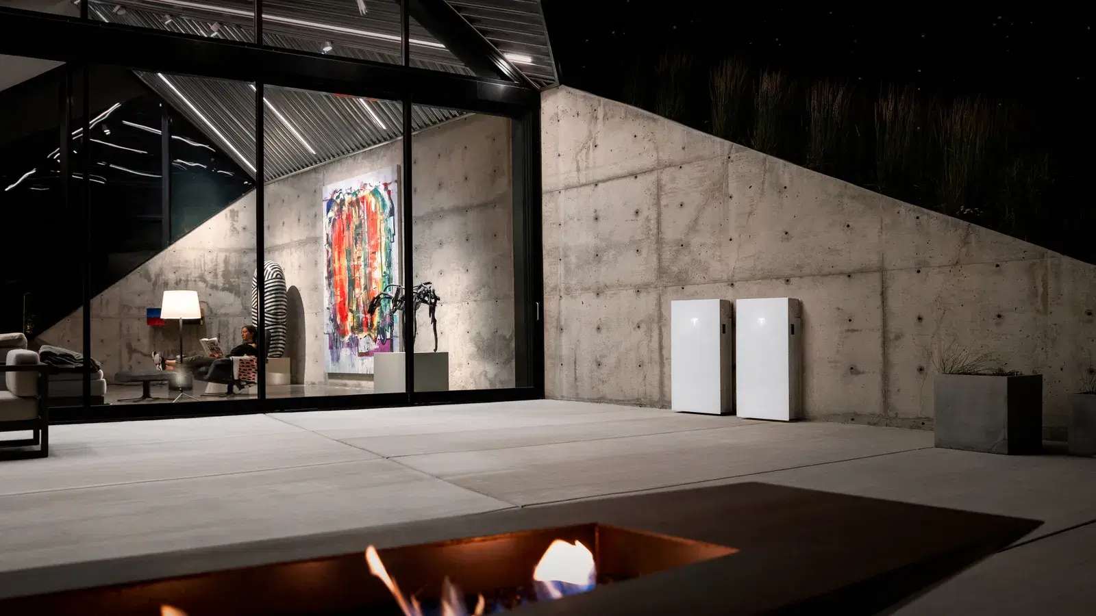 Two white Tesla Powerwalls mounted on a concrete wall on a modern patio at night, with a fire pit in the foreground and a lit glass-walled living room behind.