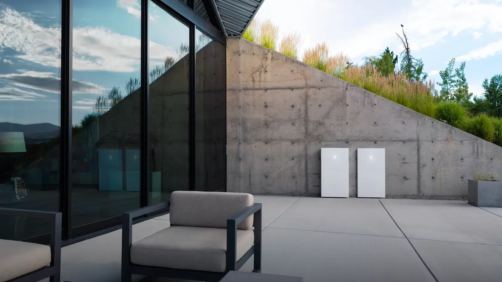 Two white Tesla Powerwalls mounted on a concrete wall on a daylight patio with modern lounge chairs and a green planted roof.
