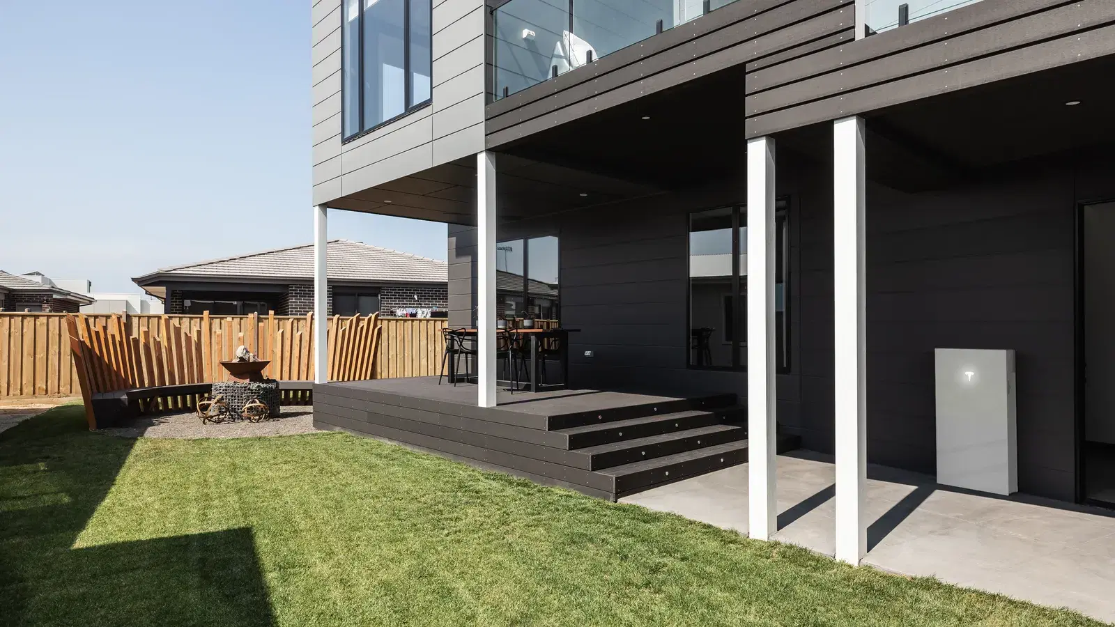 A white Tesla Powerwall mounted on the exterior wall of a contemporary two-story home with dark cladding, beside a wooden deck and lawn.