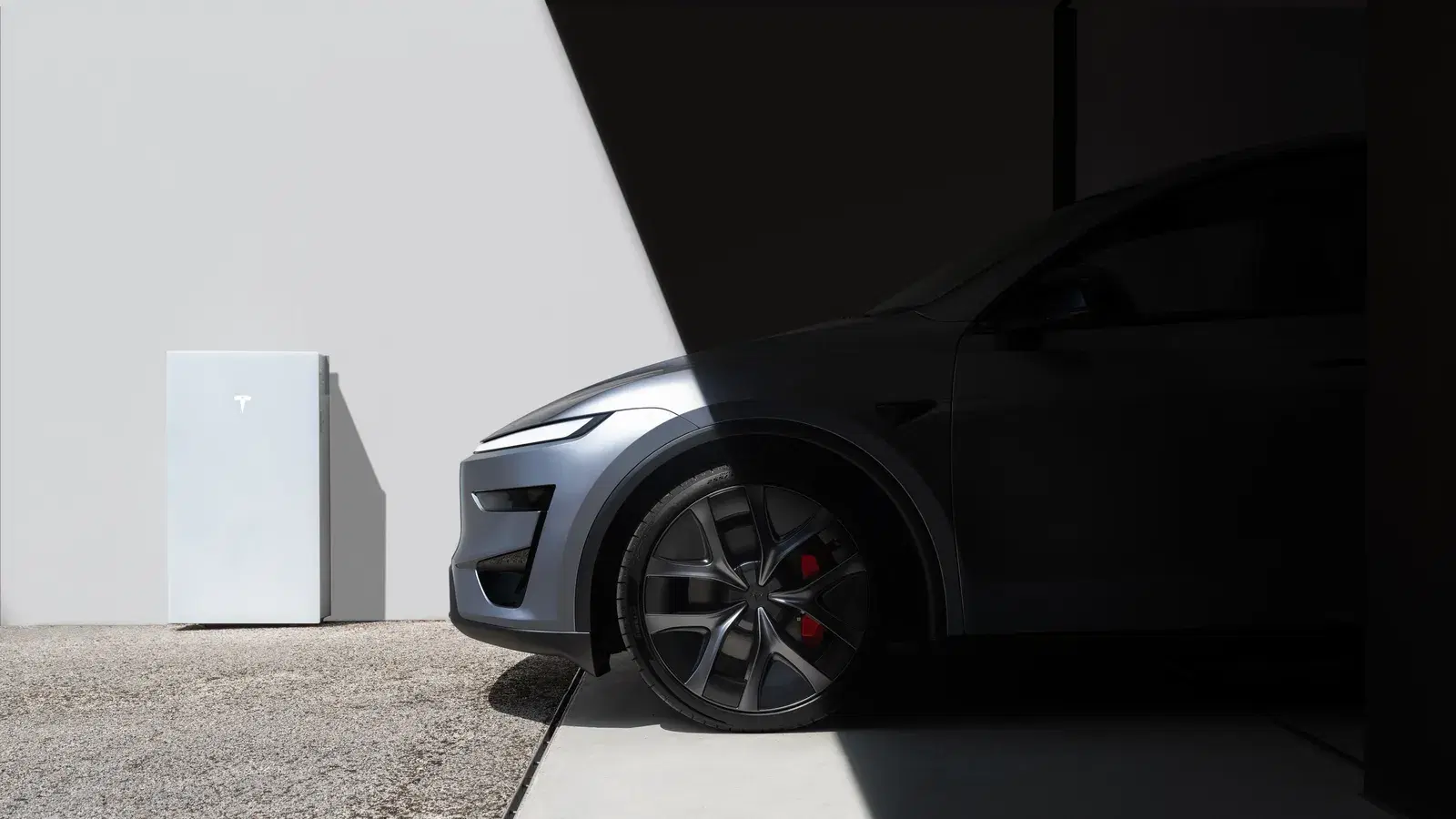 A white Tesla Powerwall on a wall beside the front wheel and headlight of a gray Tesla SUV, half in bright sun and half in shadow.