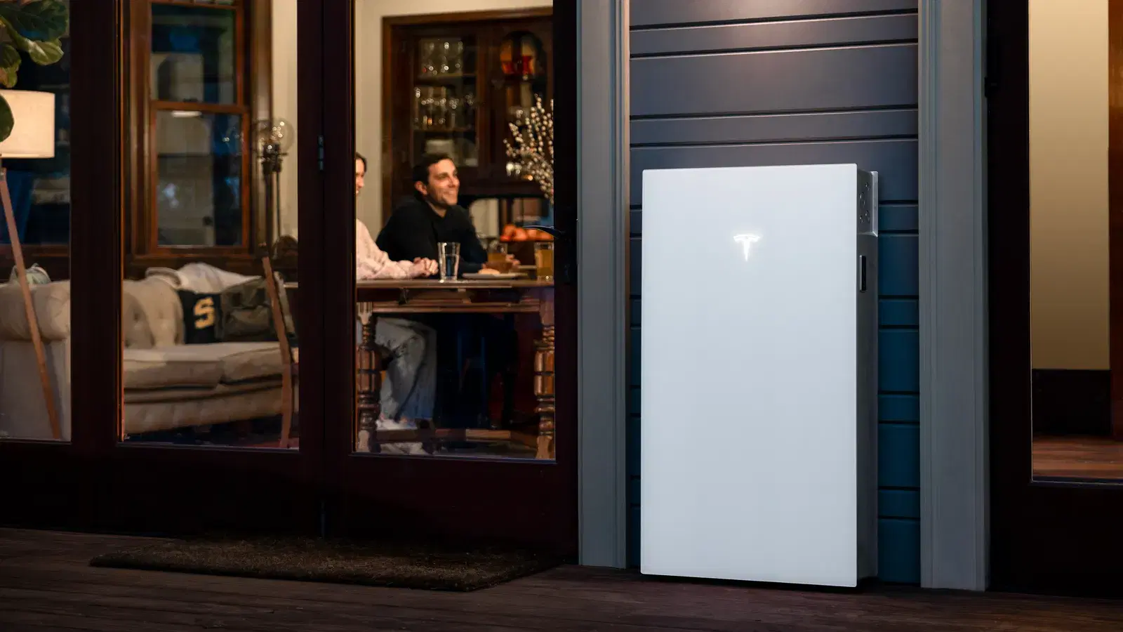 White Tesla Powerwall battery mounted on an exterior wall at twilight, with a family dining visible through the window.