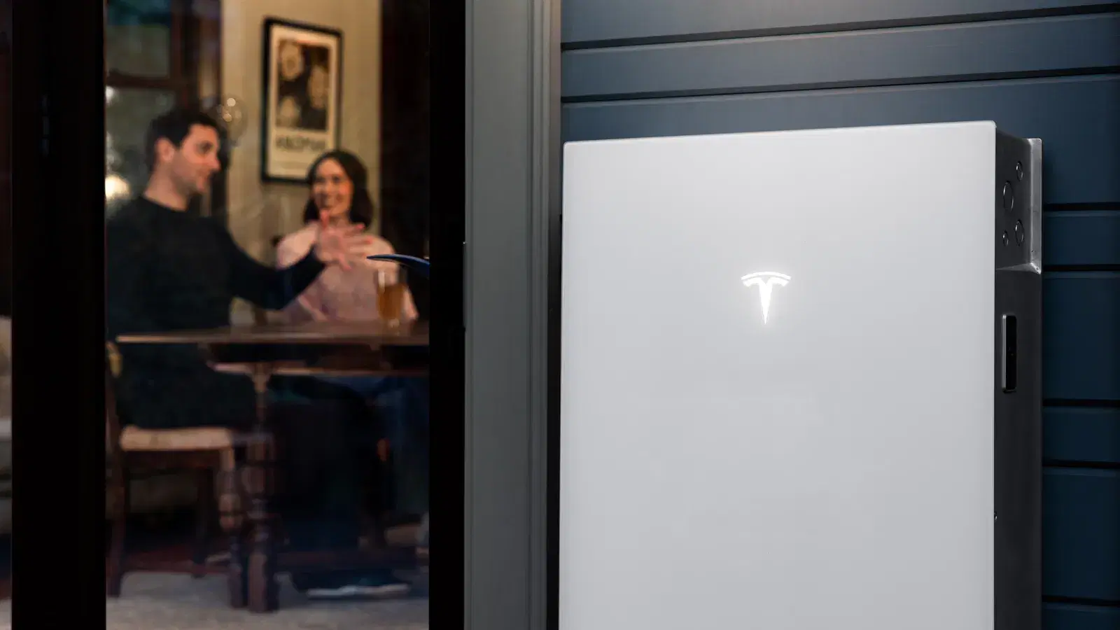 Close-up of a Tesla Powerwall with an illuminated logo, a couple raising glasses through the window behind it.
