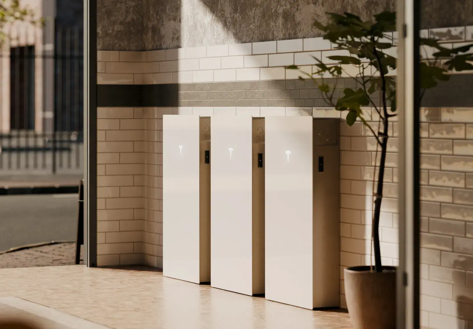 Three white Tesla Powerwall batteries installed side by side against a tiled wall, beside a potted plant.