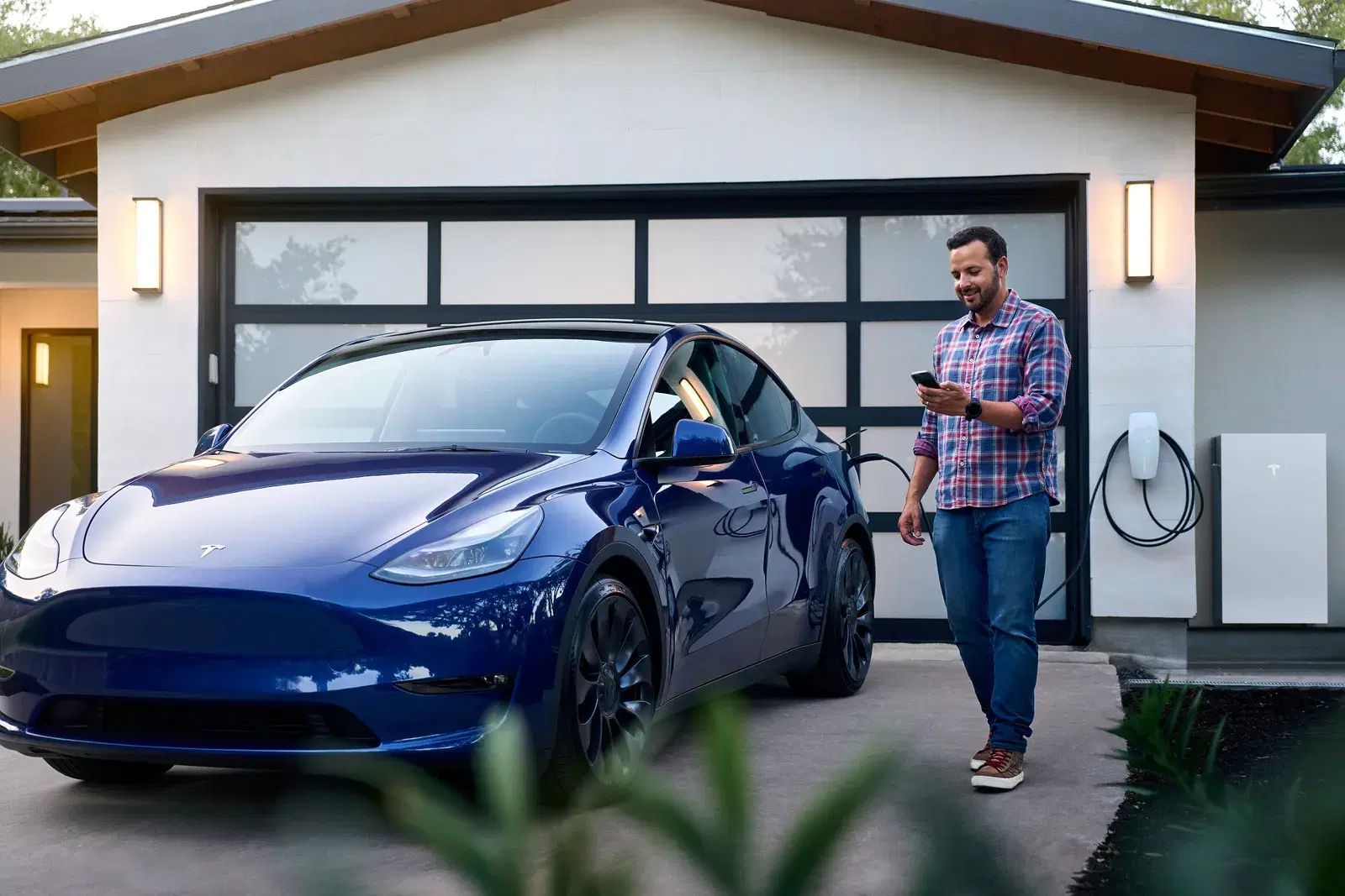 A man in a plaid shirt looks at his phone next to a blue Tesla Model Y, with a Tesla Powerwall and charger on the garage wall.