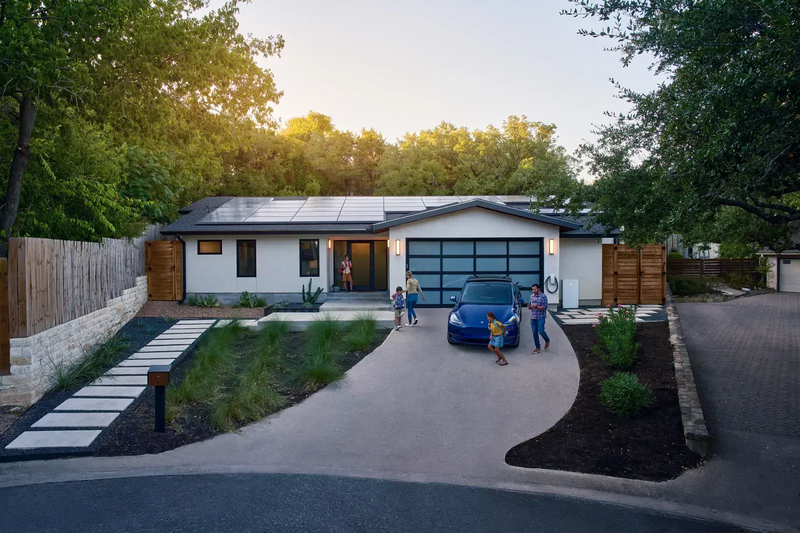 A family stands in the driveway of a single-story home at dusk with rooftop solar, a blue Tesla, and a Tesla Powerwall on the garage.