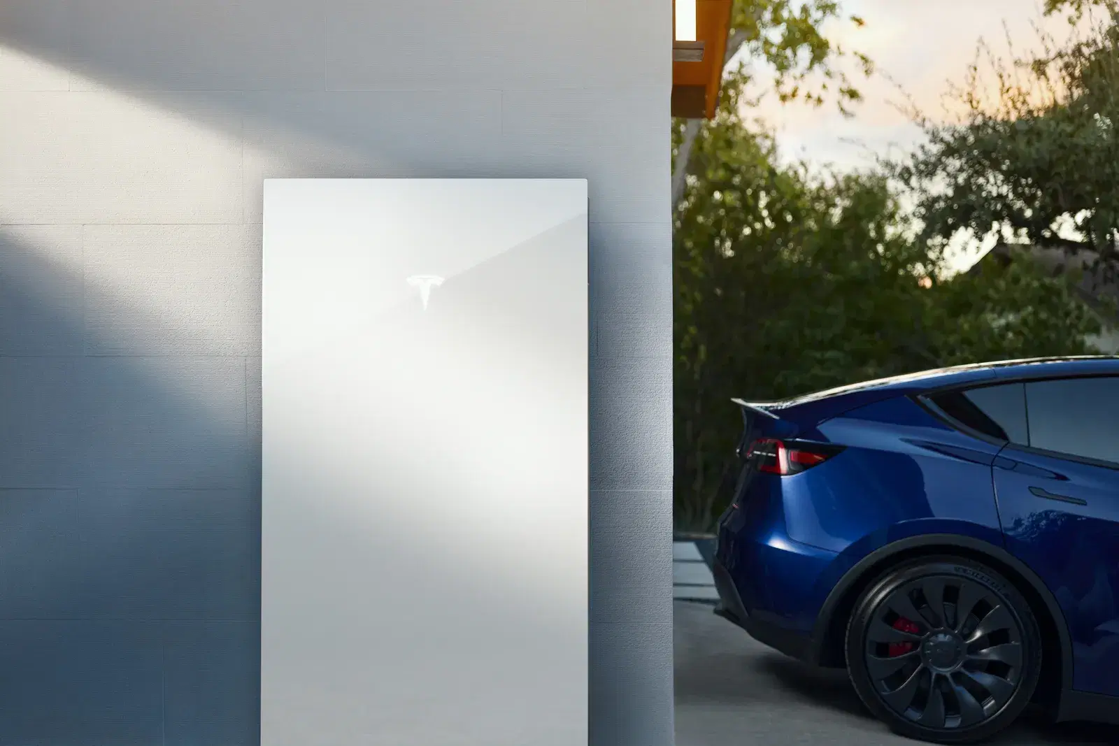 Close-up of a white Tesla Powerwall mounted on a wall with a blue Tesla Model Y parked beside it in warm evening light.