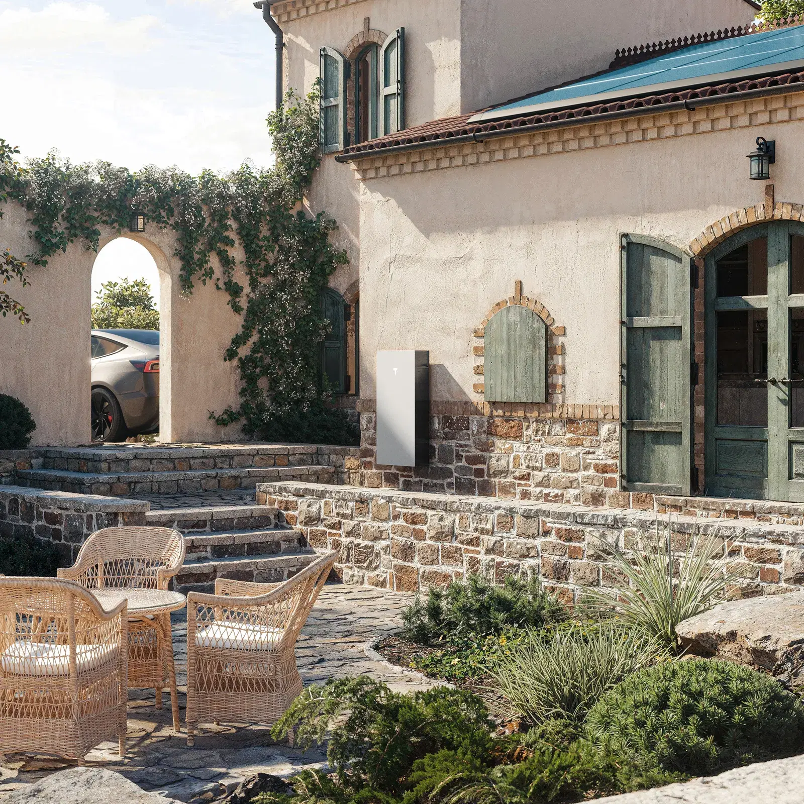 White Tesla Powerwall battery on a stone-walled courtyard of a Mediterranean-style home, wicker chairs in the foreground and a car under an arch.