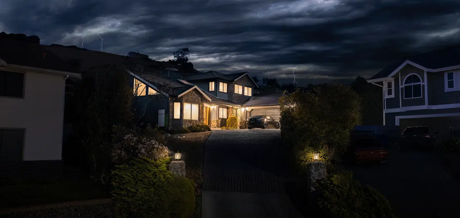 Dramatic night scene of a lit stone home under a stormy lightning sky, with a Tesla Powerwall on the exterior wall and a car in the driveway.