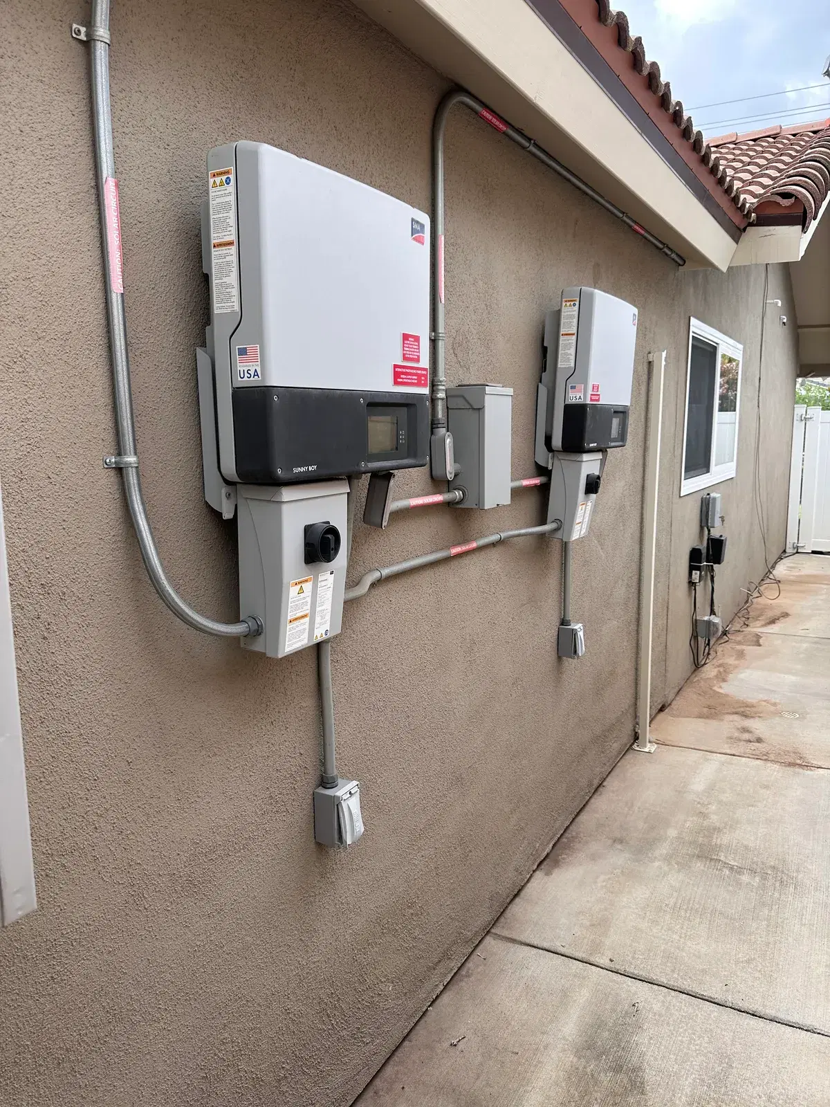 Two grey solar string inverters connected by metal conduit and disconnect boxes on a flat rooftop deck.