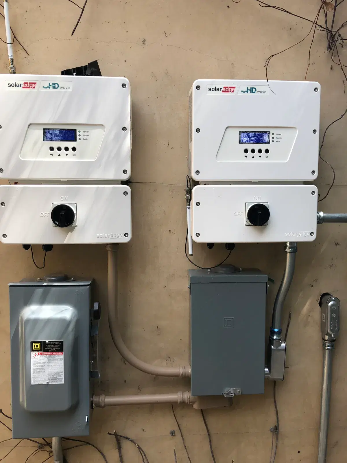 Two white SolarEdge HD-Wave string inverters with display screens, mounted beside gray AC and DC safety disconnect switches and conduit on a wall.