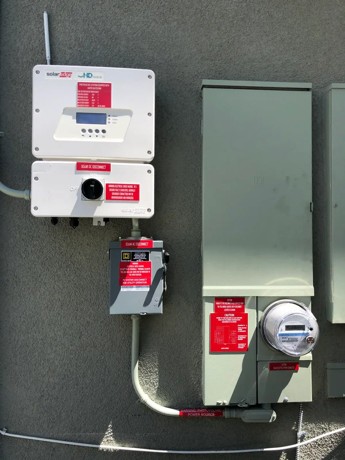 Top-down view of solar electrical equipment including a SolarEdge inverter, disconnect switches, and a utility meter mounted on a wall.