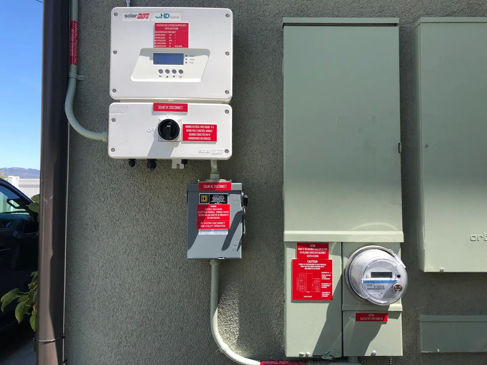 Exterior stucco wall with a SolarEdge inverter, DC and AC disconnect boxes, electrical panels, and a utility meter connected by conduit.