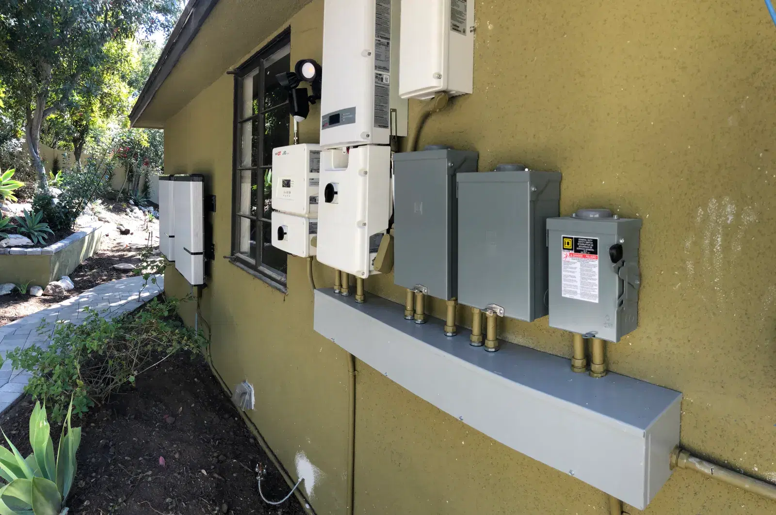 Exterior home wall with a mounted string inverter, several gray electrical disconnect boxes, and sub-panels connected by metal conduit, with no people present.