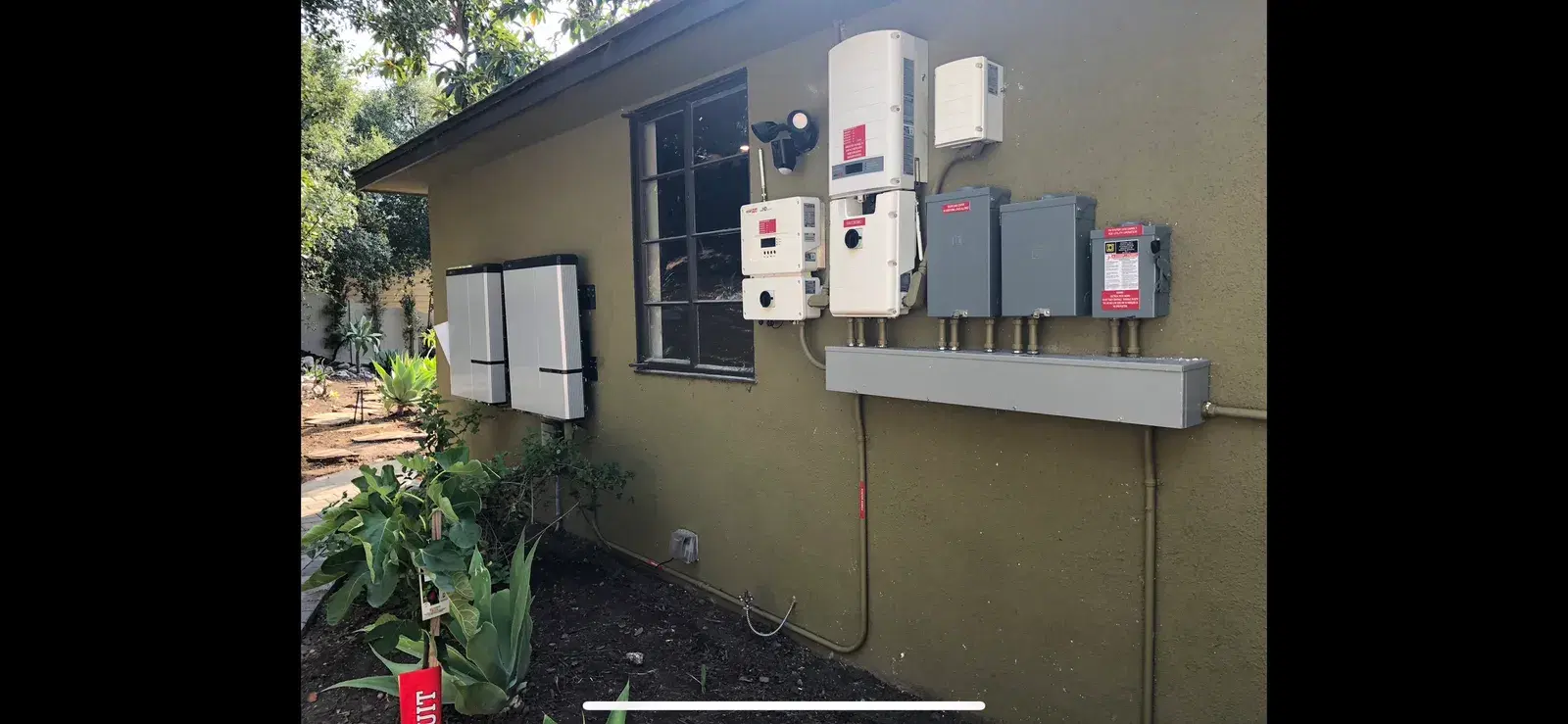 Exterior home wall mounted with solar PV electrical equipment including two string inverters, AC and DC disconnects, electrical sub-panels, and a metal wireway with conduit.
