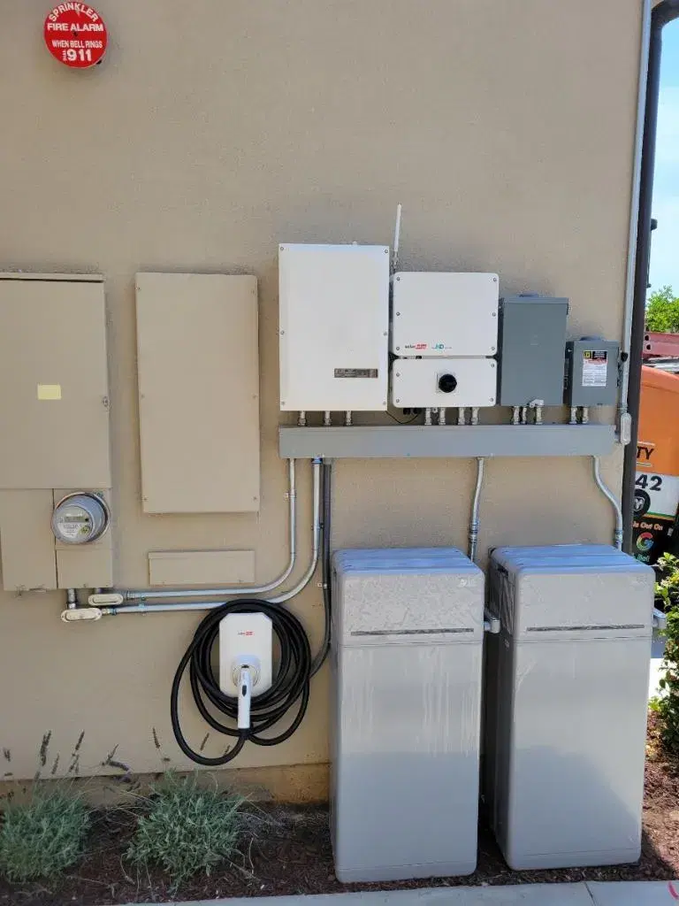 Exterior wall with a SolarEdge inverter, electrical sub-panels, conduit, an EV charger, and two gray equipment tanks below.