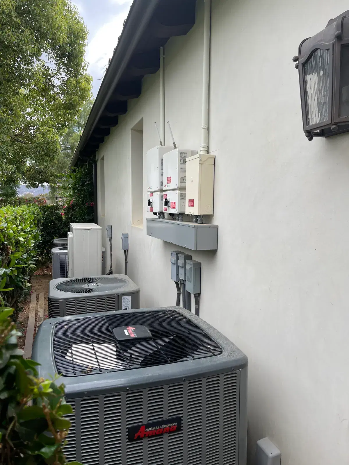 Electrical disconnects and panels on a stucco wall beside an Amana air-conditioning condenser unit outdoors.