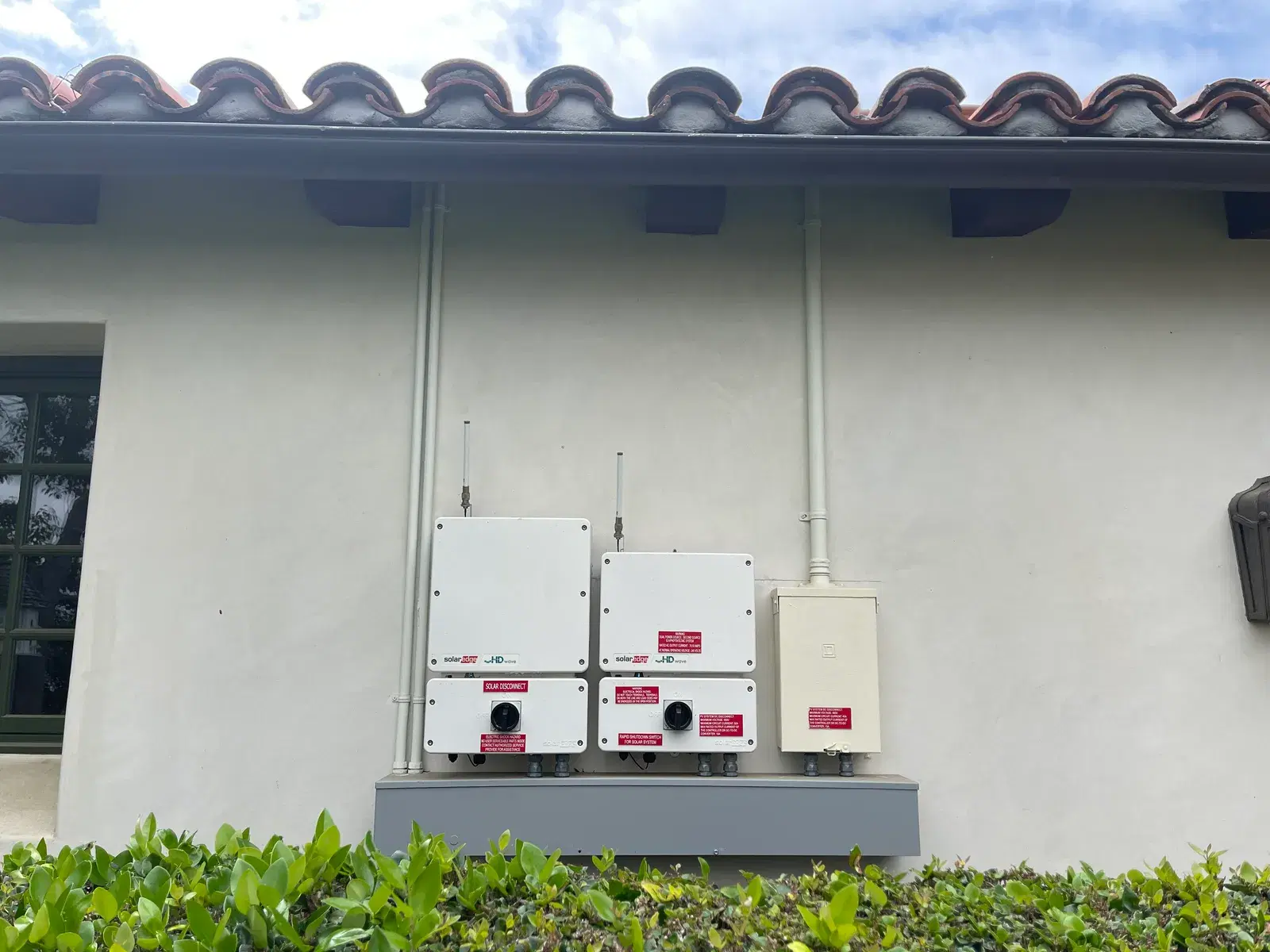 Two white SolarEdge inverters with solar disconnect boxes mounted on a stucco wall under a red clay tile roof.
