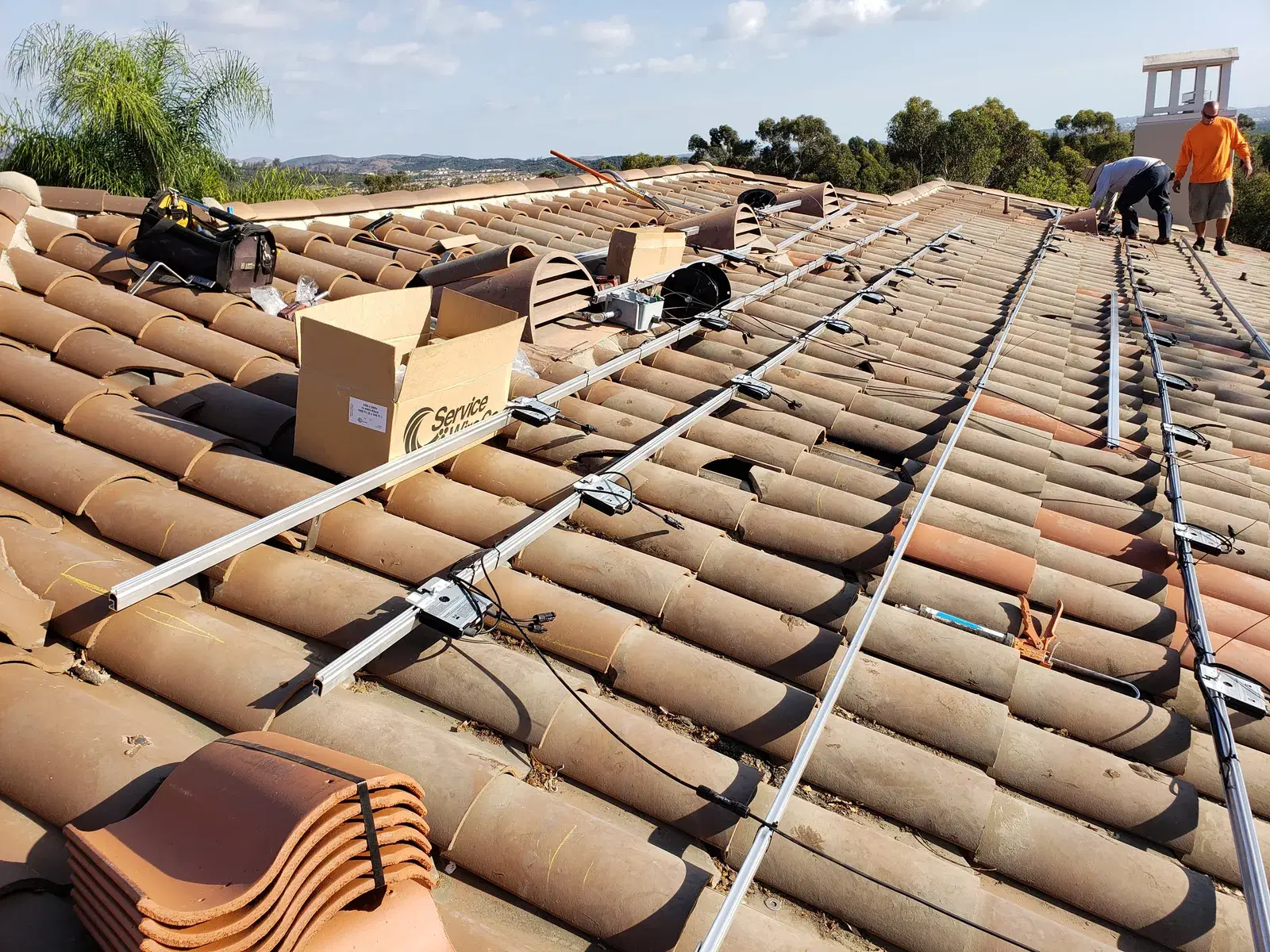 Solar installers working on a clay tile roof with mounting rails, standoffs, and conduit laid out mid-installation, with a worker in an orange shirt in the background.