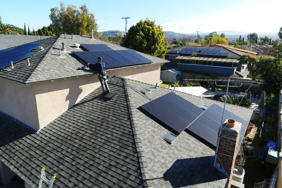 A solar technician stands on a shingle roof tending to a row of black solar panels, with suburban rooftops and hills in the background.