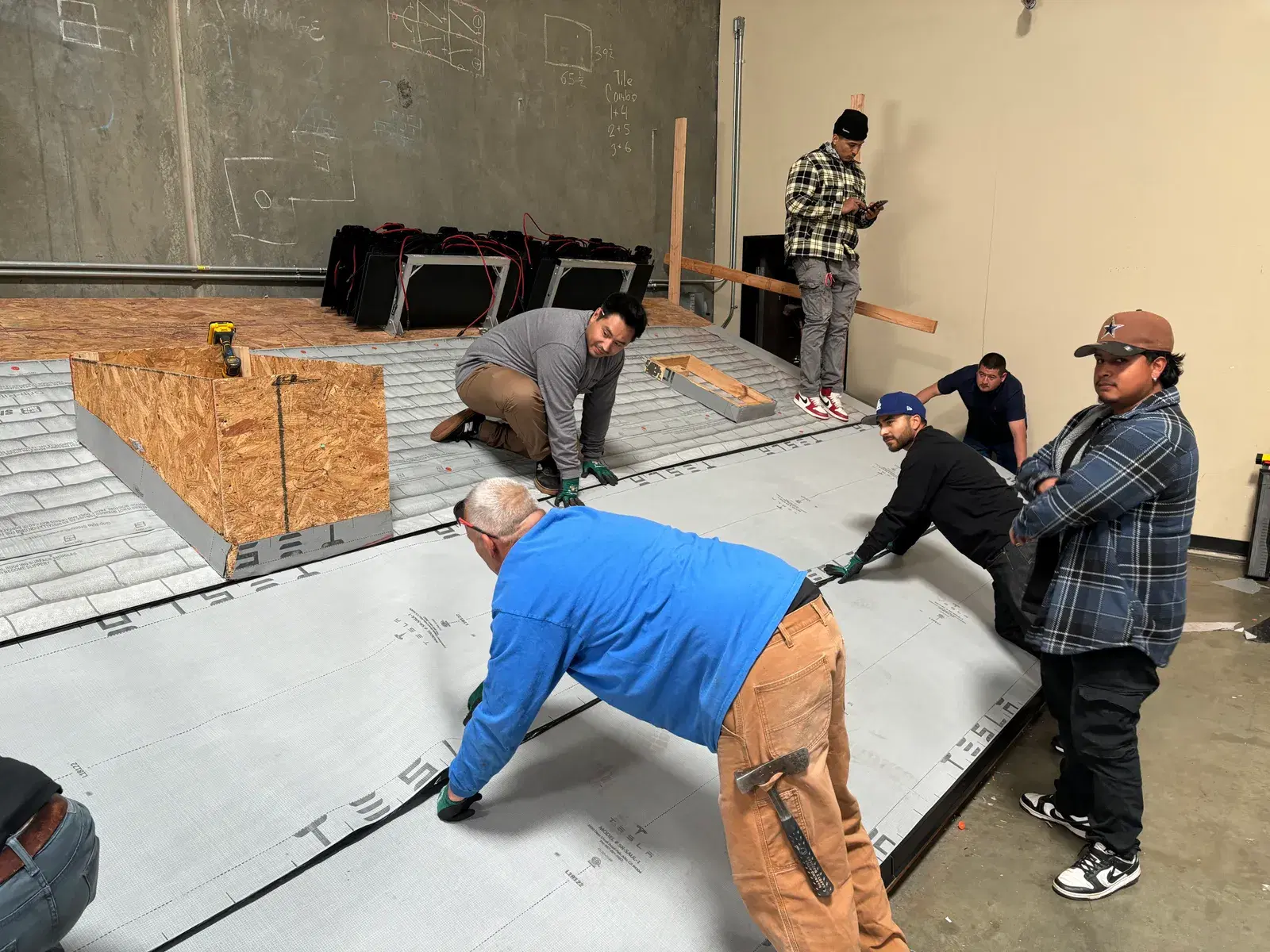 Several OC Solar workers kneel and lean over large white Tesla-branded panels on a warehouse floor, handling tools while battery boxes sit staged in the background.