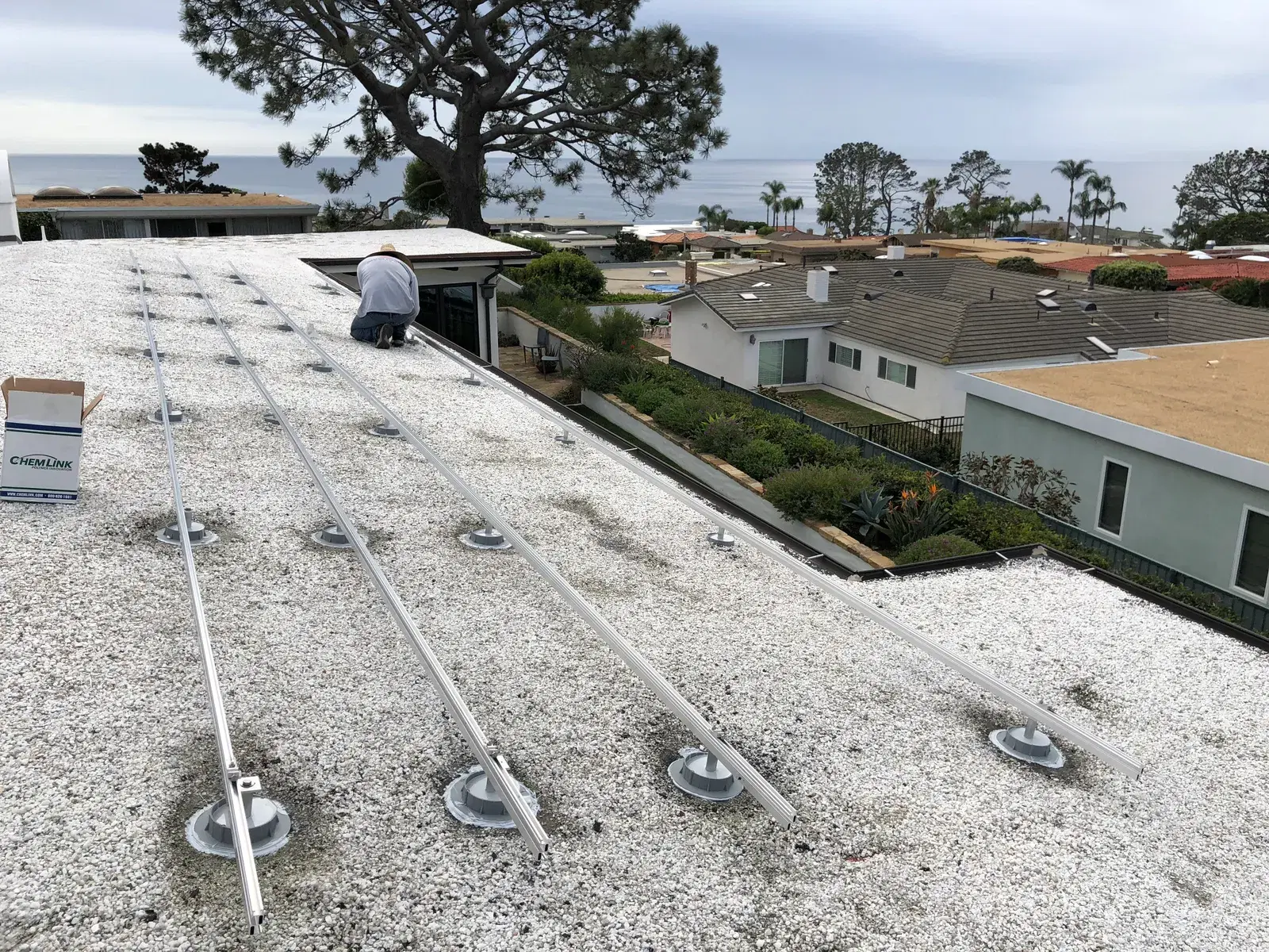 A worker crouches on a gravel rooftop installing aluminum solar racking rails with ocean and homes behind.
