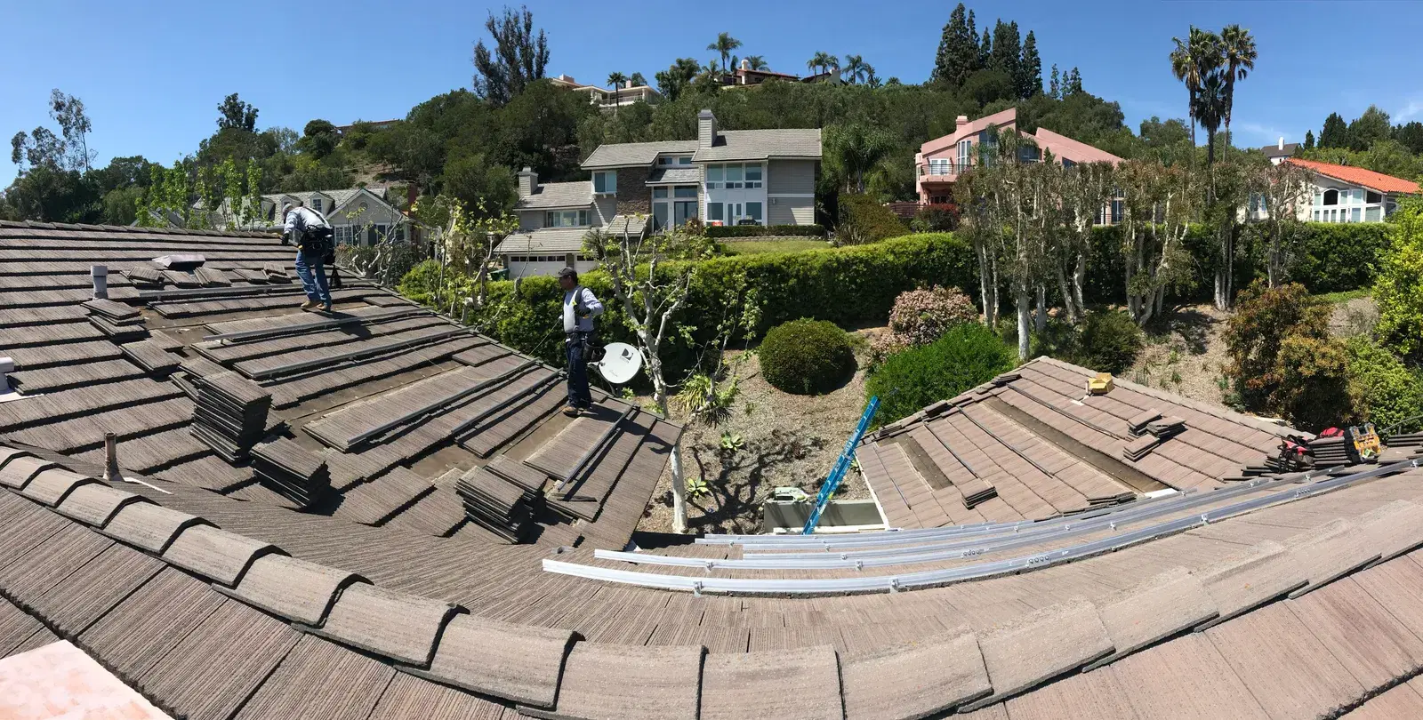 Two installers on a large concrete-tile roof laying out solar mounting rails, with hillside homes in the background.